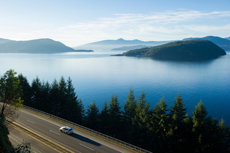 Aerial view of car driving on highway through fir trees on edge of blue lake